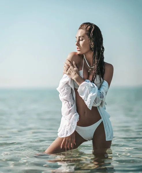 Sexy chica delgada joven en bikini blanco y camisa se encuentra en el mar tocando su hombro sensación de suavidad. Vibraciones de verano —  Fotos de Stock