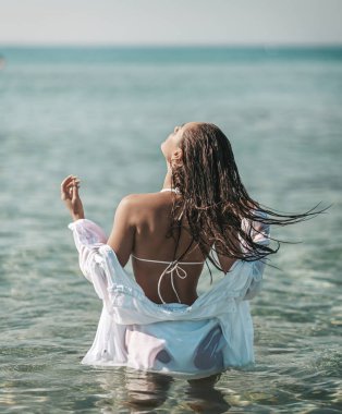 Beyaz bikinili ve tişörtlü genç seksi kadın açık deniz suyunda kameraya doğru duruyor ve başını sallıyor. Yaz titreşimleri