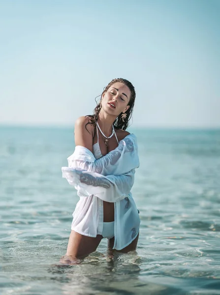 Mujer joven atractiva con gran pecho en bikini blanco y camisa se levanta en agua de mar clara abrazándose. Vibraciones de verano — Foto de Stock