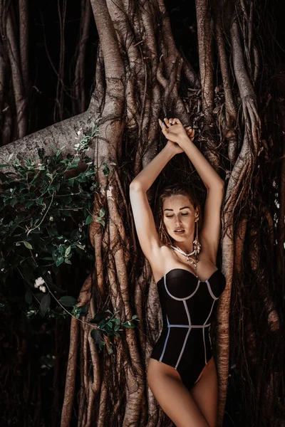 Jonge slanke vrouw in moderne bikini staat leunend naar takken van tropische bomen, sexy poserend. Zomer vibes — Stockfoto