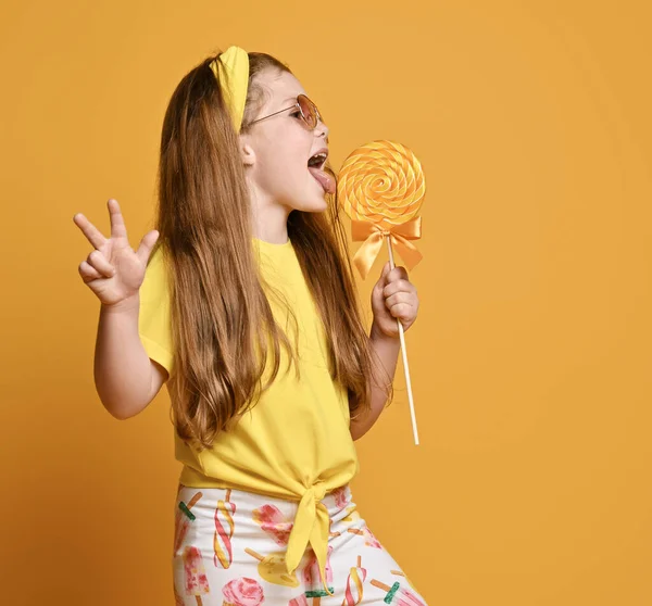 幸せな赤髪子供女の子で黄色のtシャツ,ヘッドバンドとレギンススタンド横になめる大きなロリポップキャンディー — ストック写真