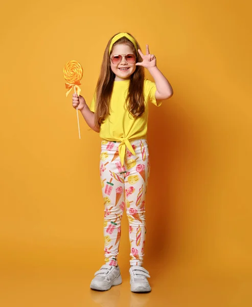 Χαριτωμένο παιχνιδιάρικο κορίτσι με κόκκινα μαλλιά σε κοριτσίστικα ρούχα κίτρινο t-shirt, κολάν και sneakers στέκεται κρατώντας μεγάλο γλειφιτζούρι καραμέλα — Φωτογραφία Αρχείου