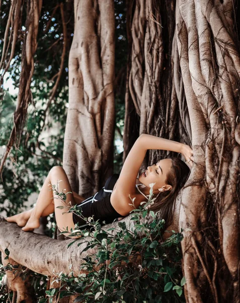 Emocionada mujer sexy en traje de baño negro está acostado en una gran rama de árbol, descansa en el bosque tropical. Vibraciones de verano — Foto de Stock