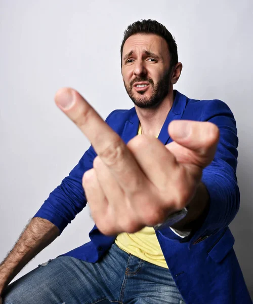 Retrato de infeliz, irritado homem adulto não raspado na jaqueta azul casual e camiseta amarela segurando o dedo médio — Fotografia de Stock