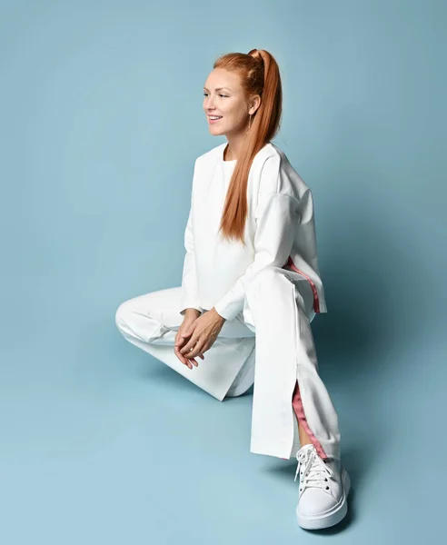 Rico ruiva jovem mulher em branco moda pantsuit com pulôver, estilo quimono senta-se em suas haunches — Fotografia de Stock