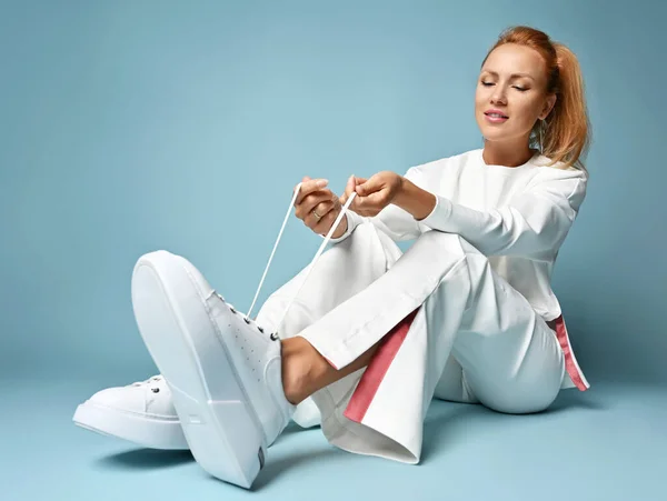 Sexy red-haired woman trendsetter in white casual pantsuit, kimono style sits on floor ties laces of her white sneakers — Stock Photo, Image