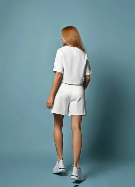 Back view. Slim woman in trendy white sport style outfit shorts and crop top, stands with her back to camera
