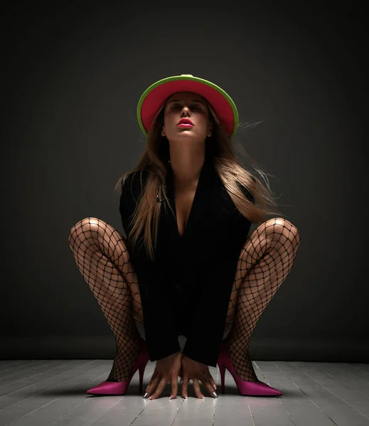 Young sexy woman in fishnet tights, pink high heels shoes and bright hat sitting on floor with legs stretched out showing lips — Stok Foto