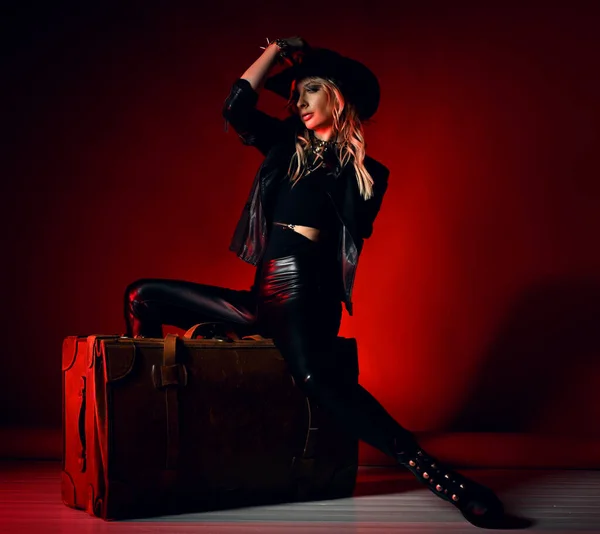 Aantrekkelijk sexy jonge blonde vrouw model in zwart lederen legging, laarzen, jas, hoed en accessoires zitten op retro stijl koffer — Stockfoto