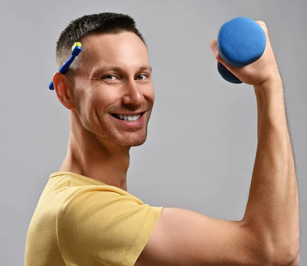 Hombre adulto alegre en camiseta amarilla sostiene cepillo de dientes detrás de su oreja y una mancuerna en la mano mira a la cámara. Vista lateral — Foto de Stock