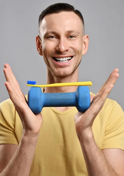 Portrait of happy friendly smiling adult man in yellow t-shirt holding toothbrush and a dumbbell with his hands — Stock Photo, Image