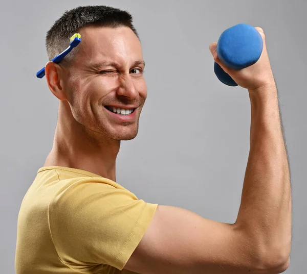 El hombre adulto sonriente con camiseta sostiene el cepillo de dientes detrás de la oreja y una mancuerna en la mano mira a la cámara y guiña los ojos. Vista lateral — Foto de Stock