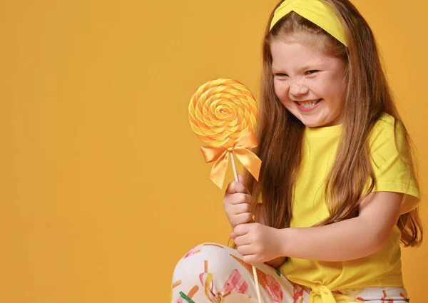 Χαρούμενο χαμογελαστό, πονηρό κορίτσι με κίτρινο μπλουζάκι και πολύχρωμα κολάν κάθεται και κοιτάζει ένα μεγάλο γλειφιτζούρι που κρατάει — Φωτογραφία Αρχείου