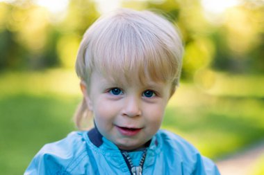 Mavi gözlü, gülümseyen ve dik bakan sarışın bir çocuğun portresi.