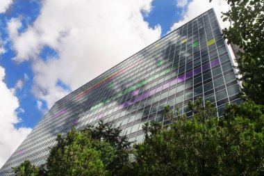 Renkli gökkuşağı ışık yansımalı cam cephe ofis binası. Etrafı yeşil ağaçlarla çevrili. Mavi bulutlu gökyüzüne karşı.