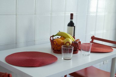 İki kişilik parlak minimalist mutfak masası, kırmızı kaplar, meyve kasesi süsü ve şarap şişesi, rahat ev yemekleri ve çağdaş yaşam tarzı konsepti.