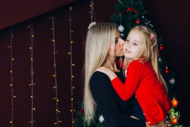 Sarışın, genç ve güzel bir anne küçük kızını bir Noel ağacının arka planında kollarında tutuyor. Yeni yılın konsepti. Tonlama.