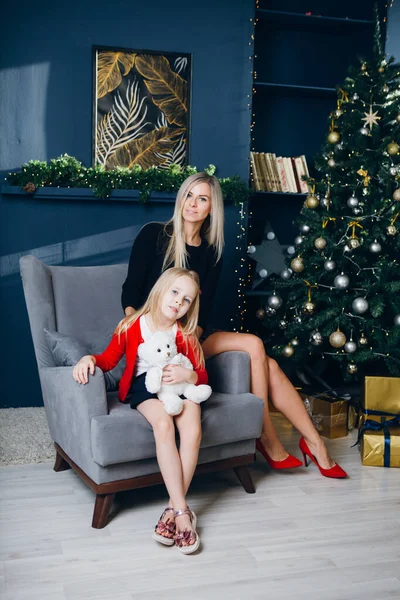 Cute young mother with blonde hair and a little daughter are sitting on a chair against the background of a Christmas tree. New year\'s concept. Toning.