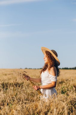 Uzun kıvırcık saçlı genç bir kız yazın gün batımında buğday tarlasında poz veriyor. Şapkalı kız. Tonlama.