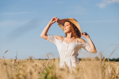 Uzun kıvırcık saçlı genç bir kız yazın gün batımında buğday tarlasında poz veriyor. Şapkalı kız. Tonlama.