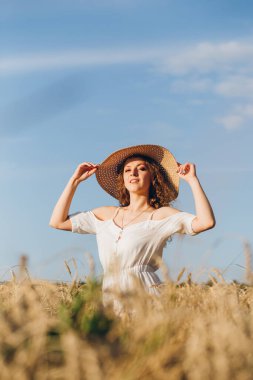 Uzun kıvırcık saçlı genç bir kız yazın gün batımında buğday tarlasında poz veriyor. Şapkalı kız. Tonlama.