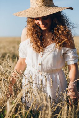 Uzun kıvırcık saçlı genç bir kız yazın gün batımında buğday tarlasında poz veriyor. Şapkalı kız. Tonlama.