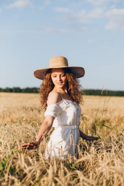 Uzun kıvırcık saçlı genç bir kız yazın gün batımında buğday tarlasında poz veriyor. Şapkalı kız. Tonlama.