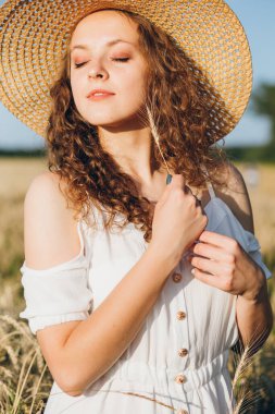 Uzun kıvırcık saçlı genç bir kız yazın gün batımında buğday tarlasında poz veriyor. Şapkalı kız. Tonlama.