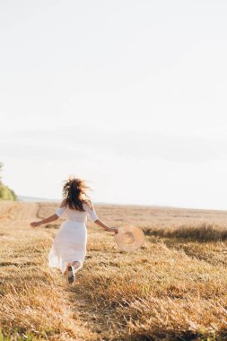 Uzun kıvırcık saçlı genç bir kız yazın gün batımında buğday tarlasında poz veriyor. Kız tarla boyunca yol boyunca koşar. Arkadan manzara. Tonlama.