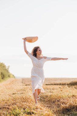 Uzun kıvırcık saçlı genç bir kız yazın gün batımında buğday tarlasında poz veriyor. Kız tarla boyunca yol boyunca koşar. Tonlama.