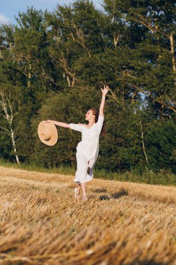 Uzun kıvırcık saçlı genç bir kız yazın gün batımında buğday tarlasında poz veriyor. Kız tarla boyunca yol boyunca koşar. Tonlama.