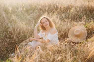 Uzun kıvırcık saçlı genç bir kız yazın gün batımında buğday tarlasında poz veriyor. Tonlama.