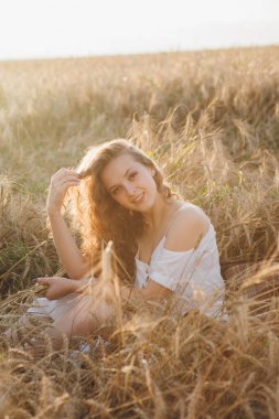 Uzun kıvırcık saçlı genç bir kız yazın gün batımında buğday tarlasında poz veriyor. Tonlama.