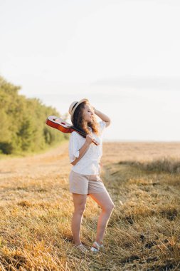 Uzun kıvırcık şortlu, genç ve güzel bir kız. Gitarlı bir şapka. Yazın gün batımında buğday tarlasında poz veriyor. Tonlama.