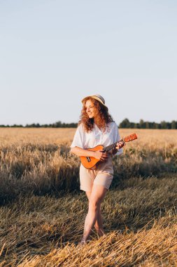 Uzun kıvırcık şortlu, genç ve güzel bir kız. Gitarlı bir şapka. Yazın gün batımında buğday tarlasında poz veriyor. Tonlama.