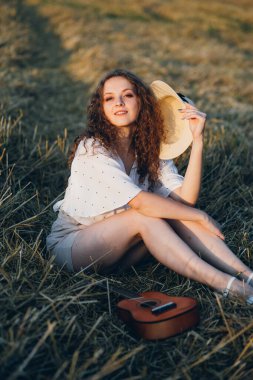 Uzun kıvırcık saçlı genç bir kız yazın gün batımında buğday tarlasında poz veriyor. Şapkalı kız. Tonlama.