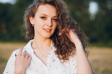 Uzun kıvırcık saçlı genç bir kız yazın gün batımında buğday tarlasında poz veriyor. Tonlama.