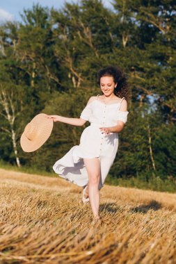 Uzun kıvırcık saçlı genç bir kız yazın gün batımında buğday tarlasında poz veriyor. Kız tarla boyunca yol boyunca koşar. Tonlama.