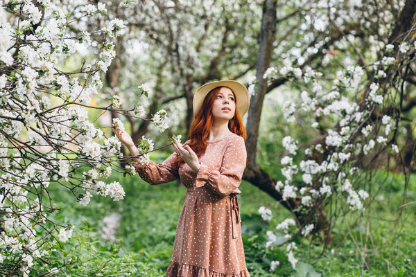 Young beautiful red-haired girl walks in a spring blooming apple orchard. Tinting.