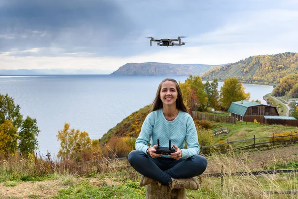 Beautiful young woman having fun with a mini drone outdoors in Lotus pose. The girl sits against the autumn forest background