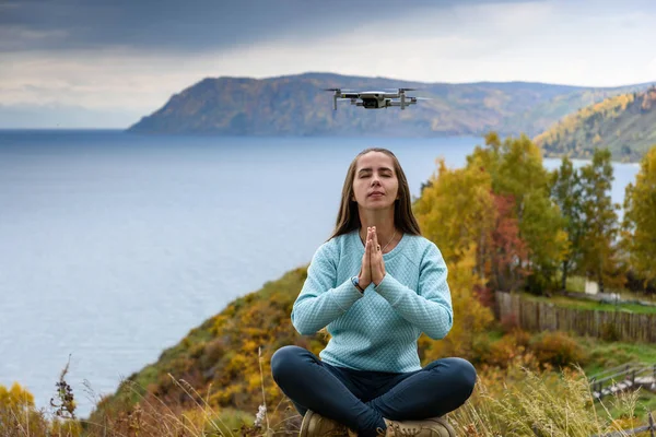 Beautiful young woman having fun with a mini drone outdoors in Lotus pose. The girl sits against the autumn forest background