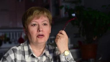 Orta yaşlı, yorgun bir kadının sağ elinde gözlük tutarak uzaklaştığı bir fotoğraf. Düşünceli bir şekilde iç çeker ve bardakları ağzına koyar.