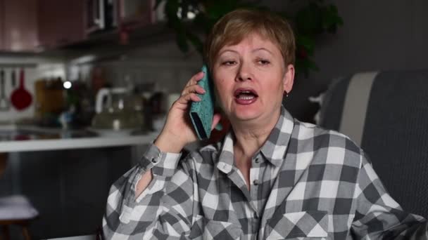 Mulher madura com cabelos castanhos sentada no sofá em casa, conversando ao telefone, acenando com a mão e sorrindo. Dentro de casa. Retrato. — Vídeo de Stock