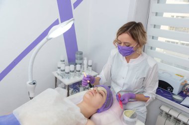Young woman cosmetologist apply green clay mask on womans face. Beautiful brunette woman in beauty salon