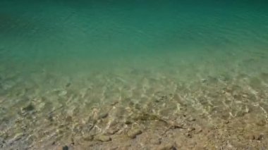 Nehirde berrak eğimli su dalgaları, suyun altında kayalık zemin. Su yüzeyinde güneş parlıyor.