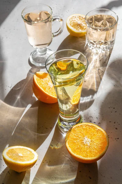 Refreshing drinks with lemon and orange in glasses in sunlight. Trendy artistic  hard shadows and rainbow highlights. Refraction effect in glass. Sliced citrus fruits on white textured background