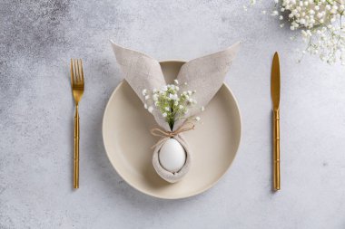 Paskalya sofrasında peçeteli yumurta, kulaklı Paskalya tavşanı, seramik tabakta küçük beyaz çiçekler ve çatal bıçak takımı. Paskalya masa süslemeleri. Üst görünüm