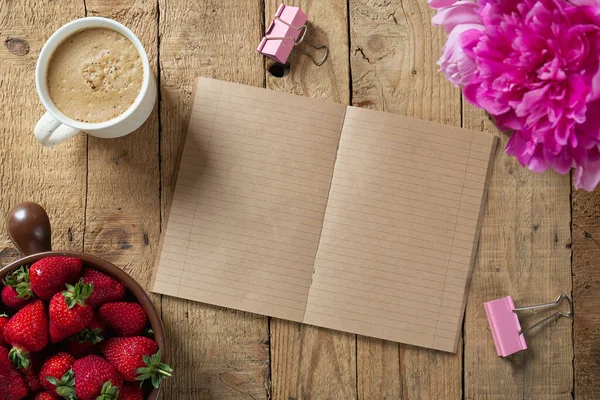 Kabaca oyulmuş ahşap bir masa, açık bir klasik defter, bir buket şakayık, bir kase çilek ve bir fincan kahve. Üst görünüm, alanı kopyala 