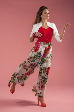 Sıska, şirin bir manken moda pozu veriyor, kırmızı pembe çiçekli bir bahar elbisesi giyiyor, kameradan uzağa bakıyor.