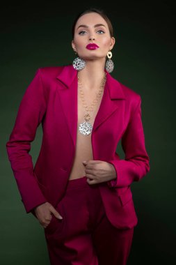 Süper model kamerada çok güzel görünüyor, çok şehvetli, zarif bir kıyafet giyiyor, fuşya takıyor, koyu yeşil arka planda poz veriyor..